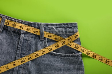 Photo of Jeans with measuring tape on green background, top view. Weight loss concept