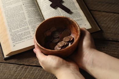 Photo of Donate and give concept. Woman holding bowl of coins, closeup. Bible and cross on wooden table
