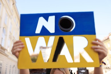 Woman holding poster in colors of Ukrainian flag with words No War near tank barrel on city street, closeup