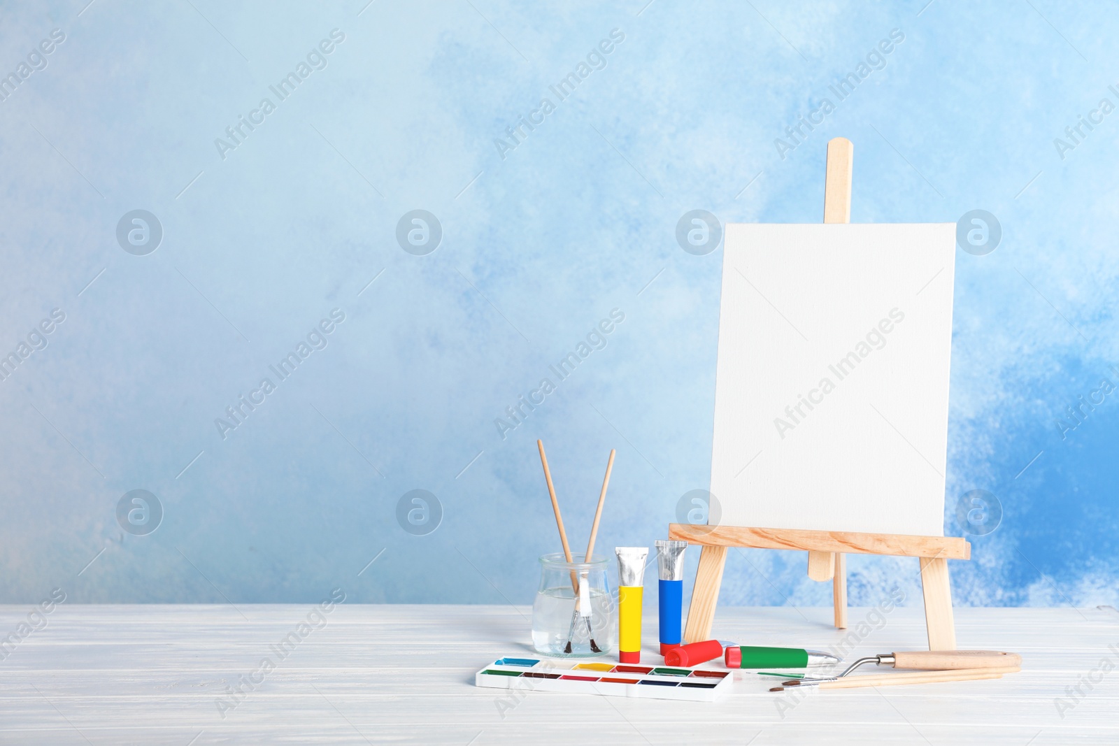 Photo of Wooden easel with blank canvas board and painting tools for children on table near color wall. Space for text