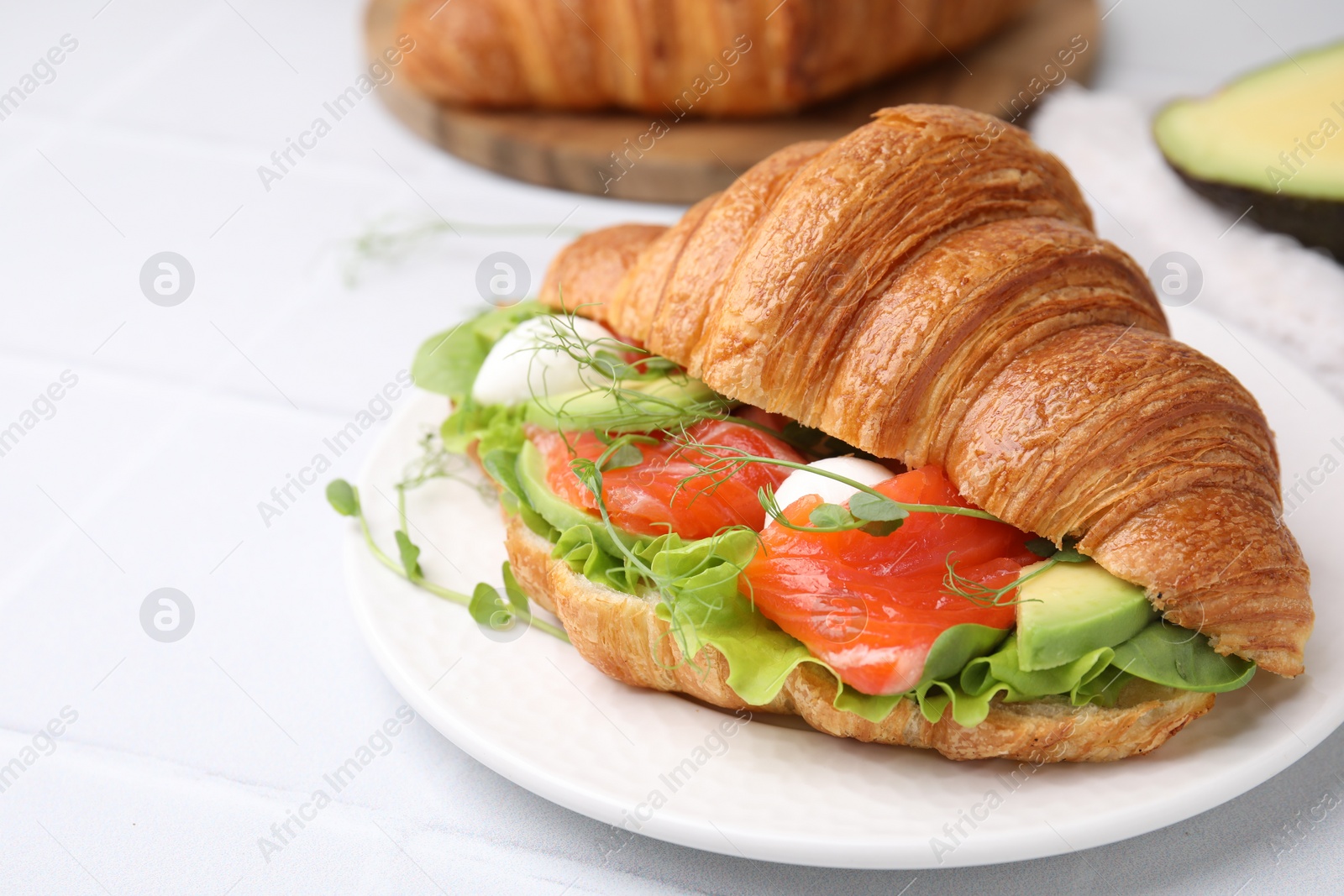 Photo of Tasty croissant with salmon, avocado, mozzarella and lettuce on white tiled table, closeup