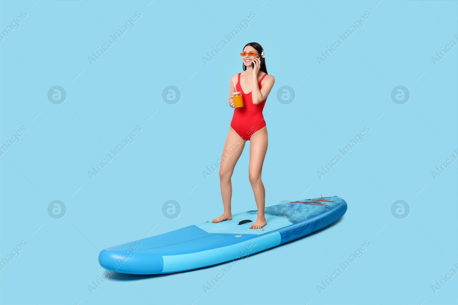 Photo of Happy woman with refreshing drink talking on smartphone on SUP board against light blue background