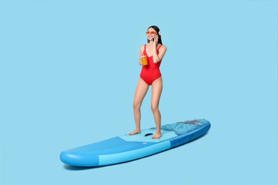 Happy woman with refreshing drink talking on smartphone on SUP board against light blue background