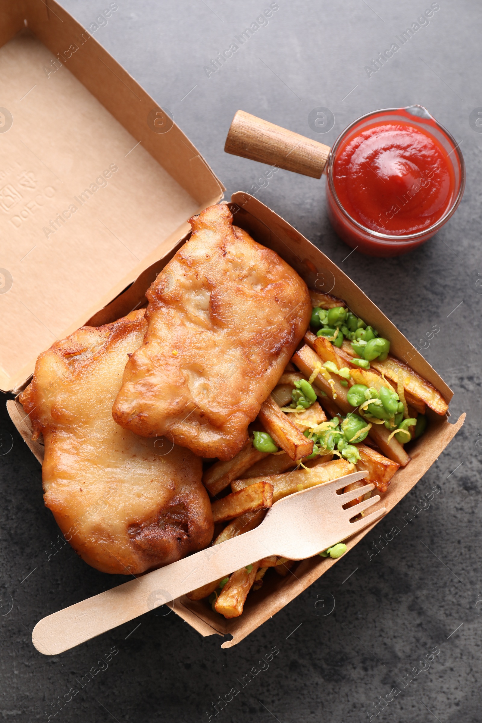 Photo of Tasty fish, chips, sauce and peas in paper box on grey table, top view