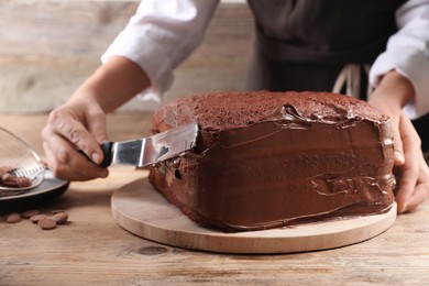 Woman smearing sides of sponge cake with chocolate cream at wooden table, closeup