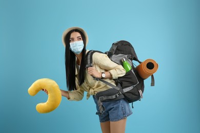 Photo of Female tourist in medical mask with backpack and travel pillow on light blue background