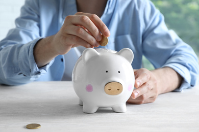 Photo of Man putting coin into piggy bank at white table, closeup