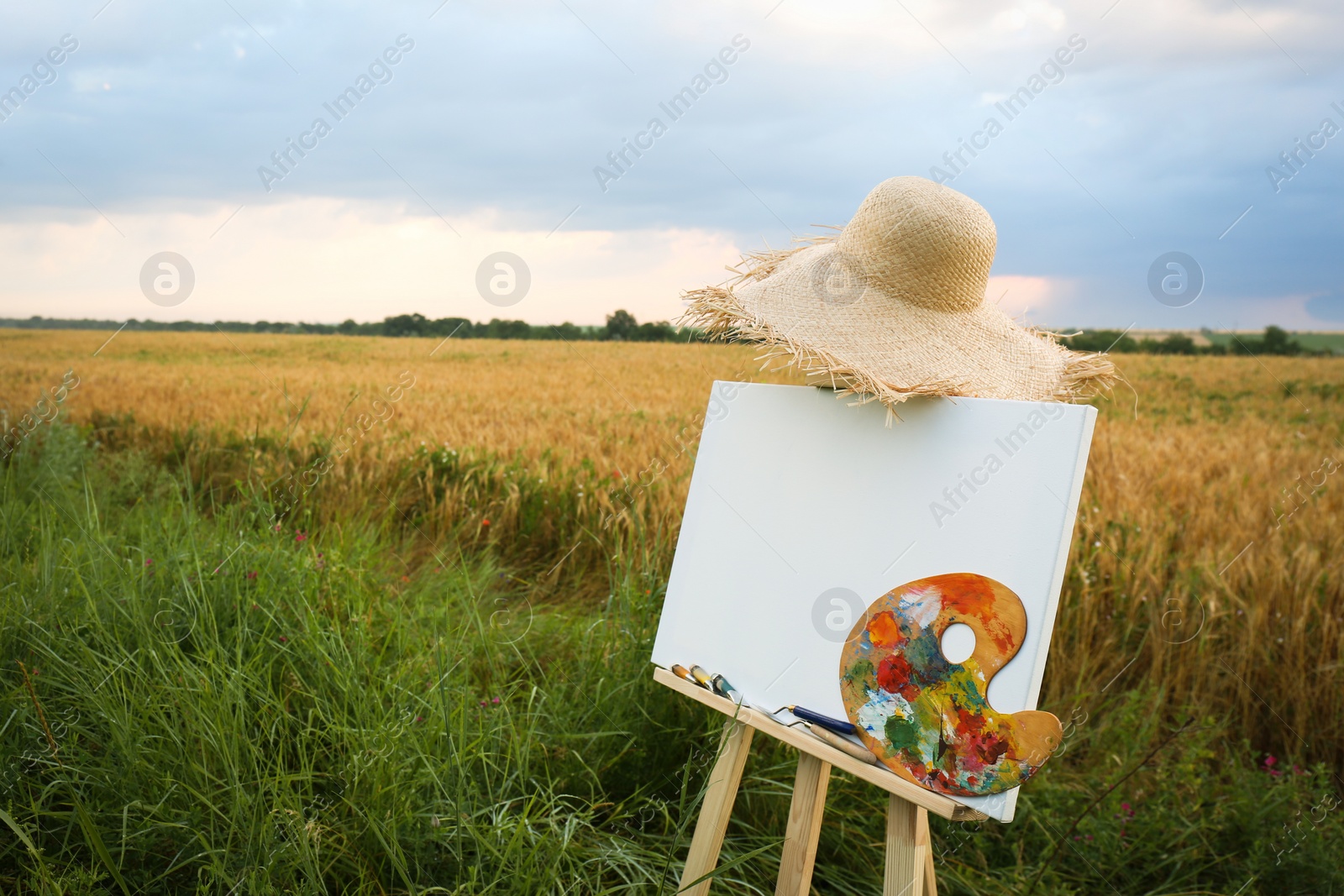 Photo of Wooden easel with blank canvas, painting equipment and hat in field. Space for text