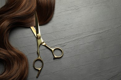 Professional hairdresser scissors with brown hair strand on dark grey table, top view. Space for text