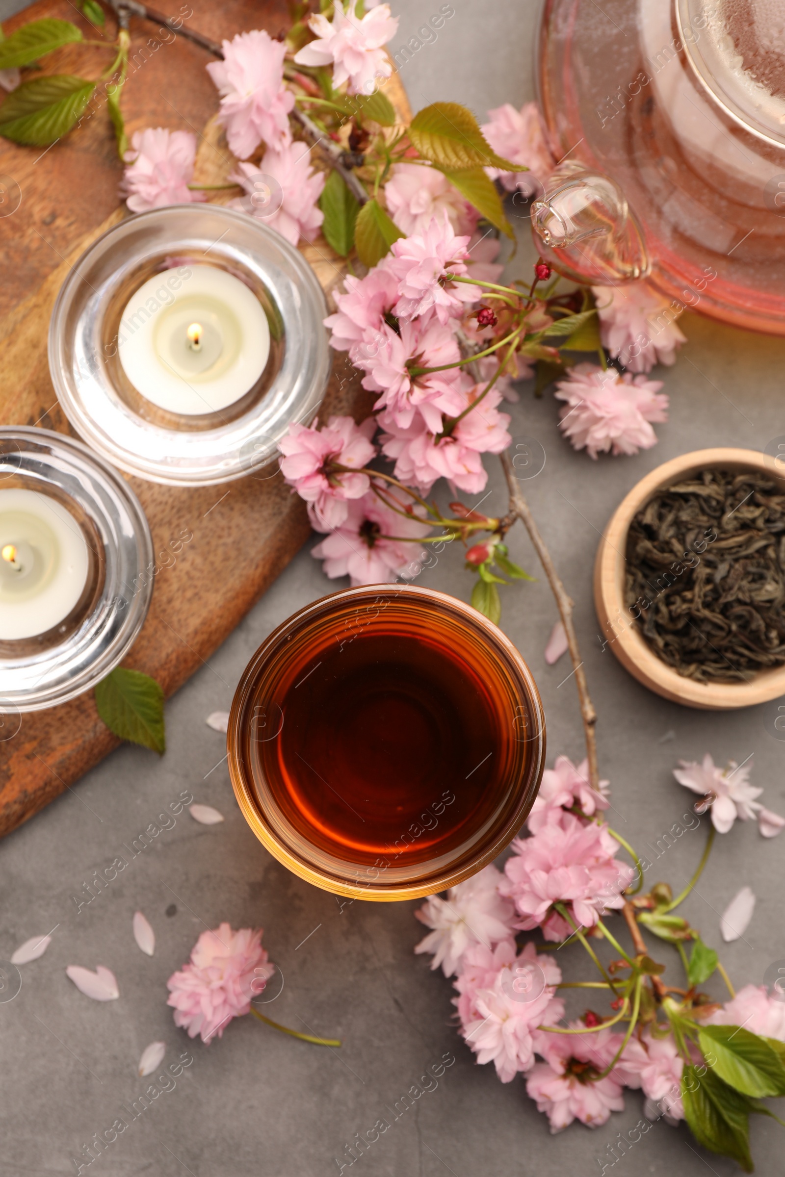 Photo of Traditional ceremony. Cup of brewed tea, burning candles, teapot and sakura flowers on grey table, flat lay