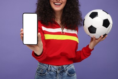Photo of Happy fan holding soccer ball and showing smartphone on violet background, closeup