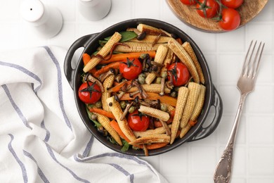 Tasty roasted baby corn with tomatoes, capers and mushrooms on white tiled table, flat lay