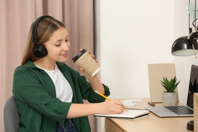 Photo of Teenage girl with headphones and cup of drink writing in notebook at wooden table indoors