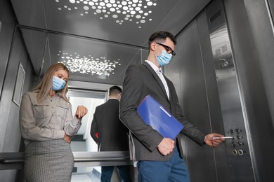 Photo of Coworkers with face masks in elevator, man using pen to choose floor, low angle view. Protective measure