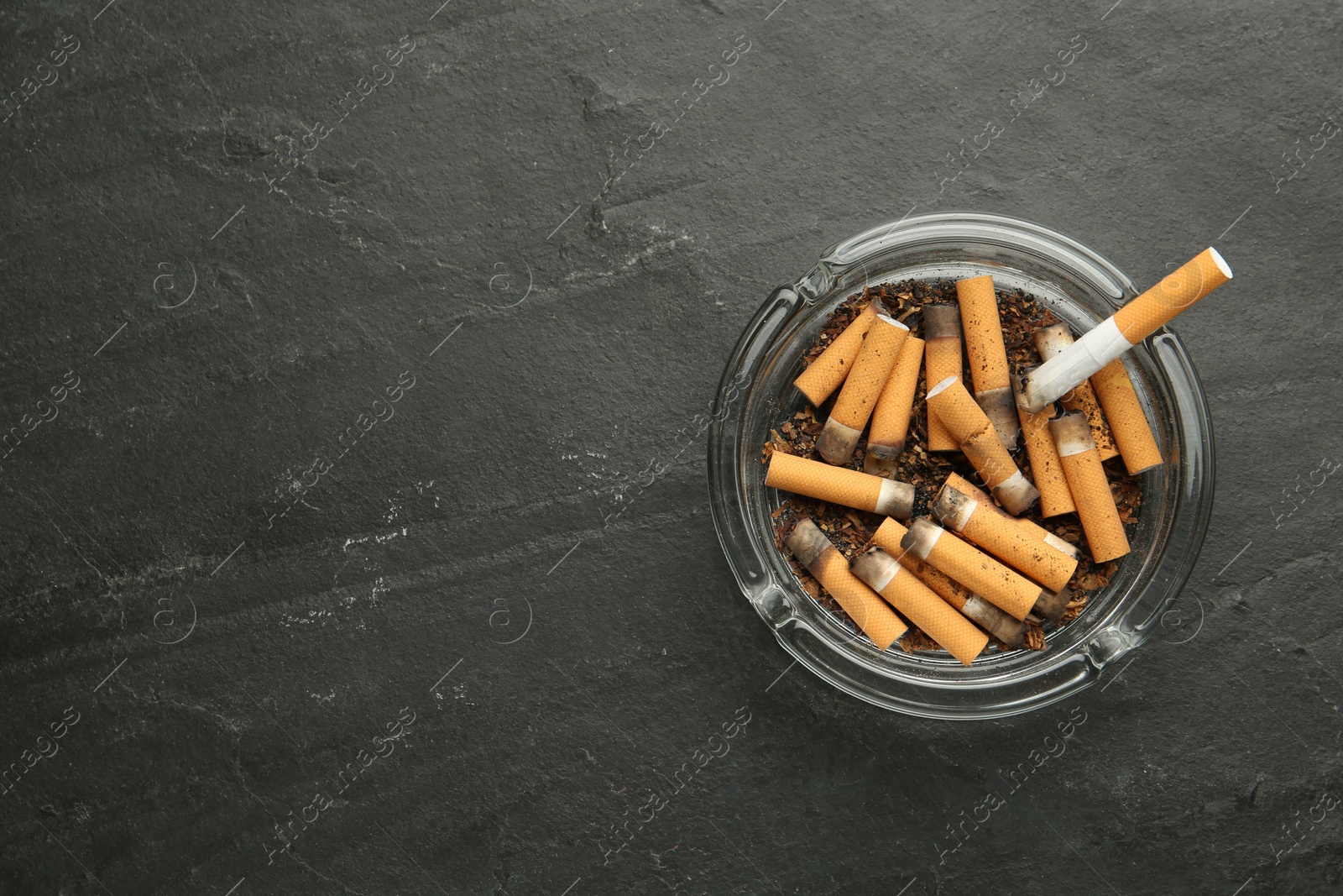 Photo of Glass ashtray with cigarette stubs on dark grey table, top view. Space for text