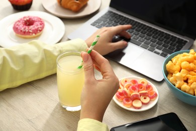 Bad eating habits. Woman with glass of drink and different snacks using laptop at wooden table, closeup