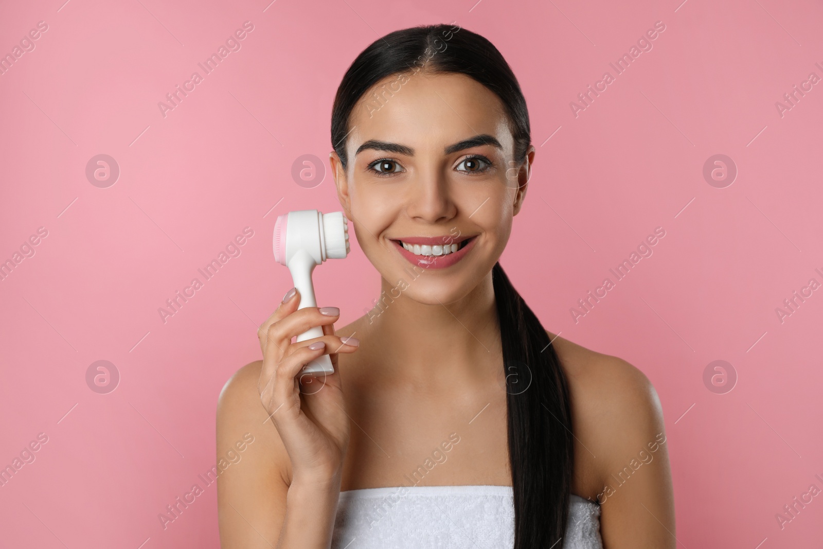 Photo of Young woman using facial cleansing brush on pink background. Washing accessory