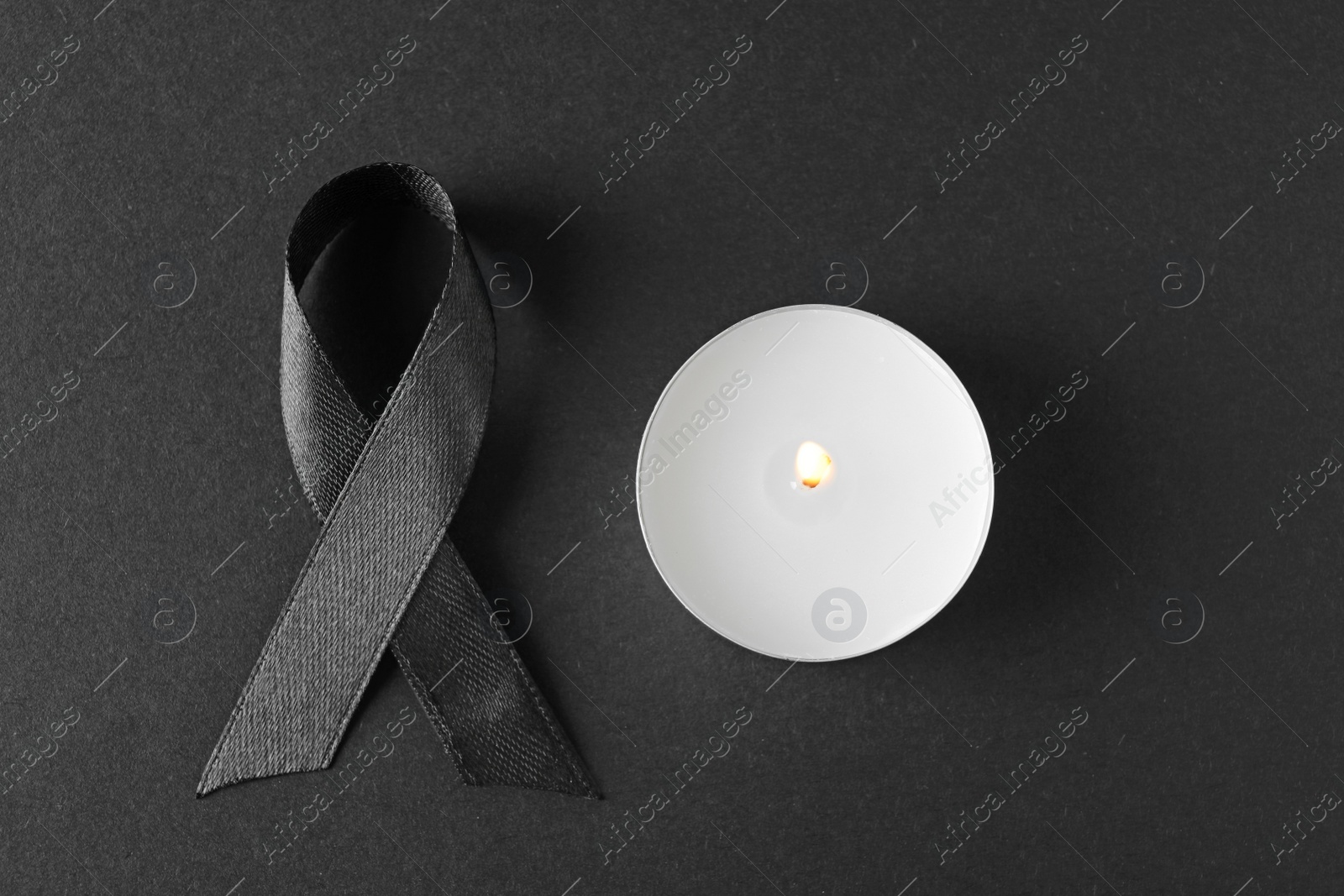 Photo of Black ribbon and burning candle on dark background, top view. Funeral symbols