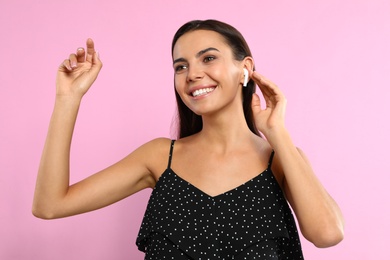 Happy young woman listening to music through wireless earphones on pink background