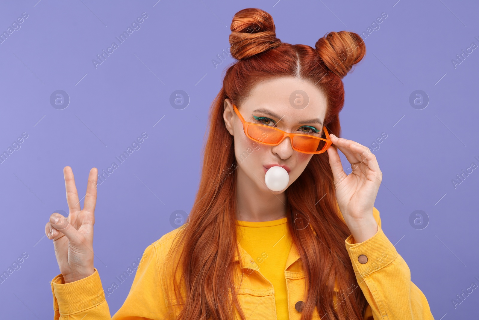 Photo of Portrait of beautiful woman in sunglasses blowing bubble gum and showing peace gesture on violet background