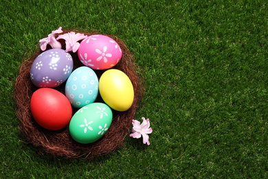 Photo of Colorful Easter eggs in decorative nest with flowers on green grass, top view. Space for text
