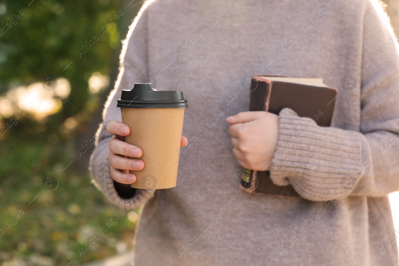 Photo of Woman holding takeaway cardboard cup and book on city street, closeup. Coffee to go