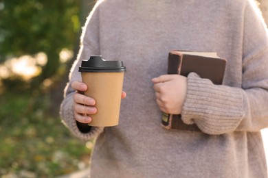 Woman holding takeaway cardboard cup and book on city street, closeup. Coffee to go