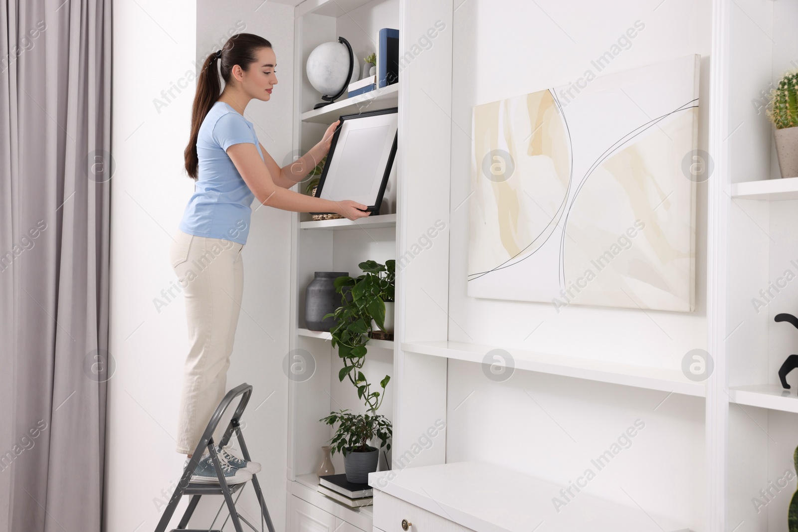 Photo of Woman on ladder with picture near shelves at home