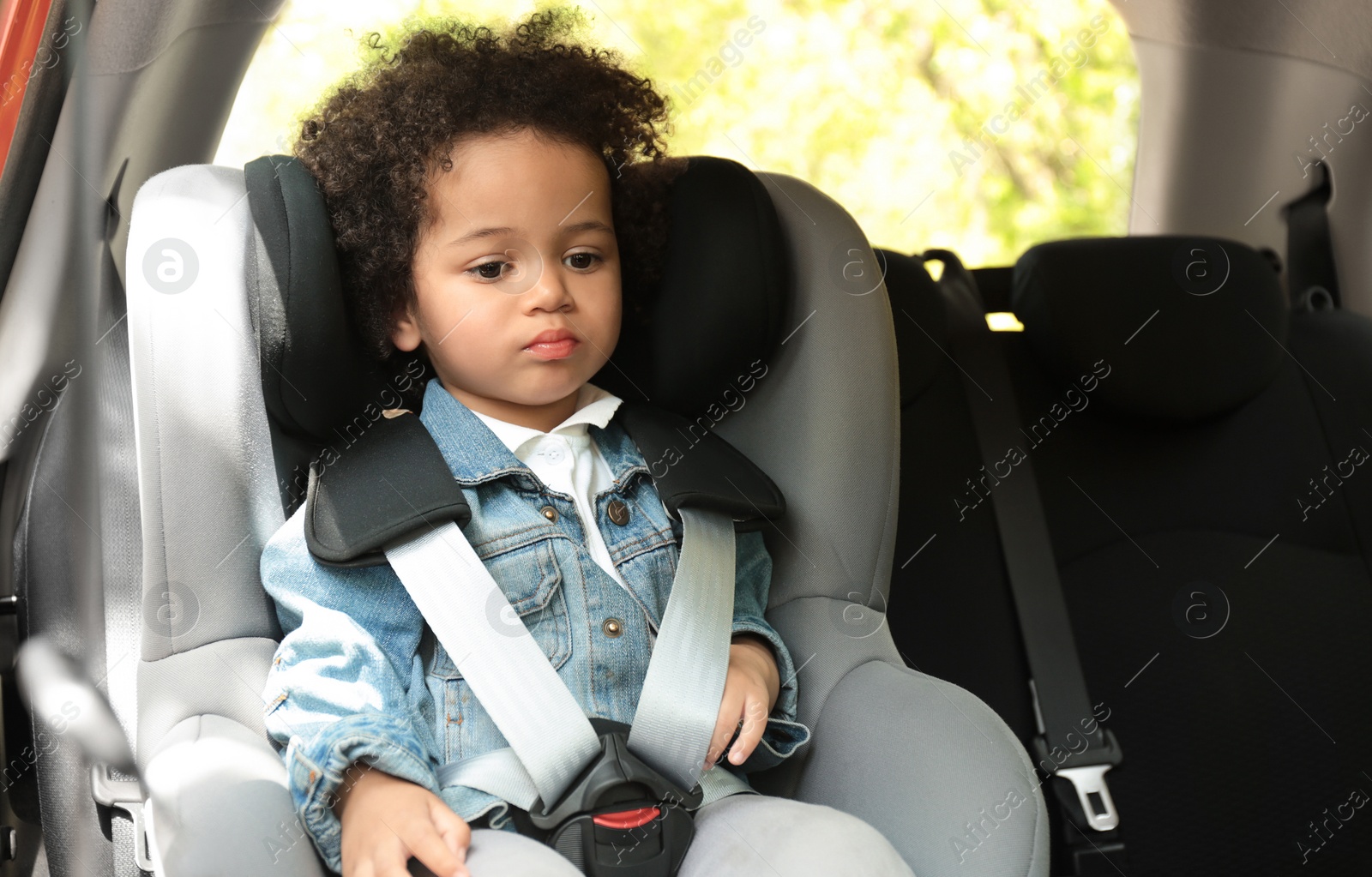 Photo of Cute African-American girl sitting in safety seat alone inside car. Child in danger