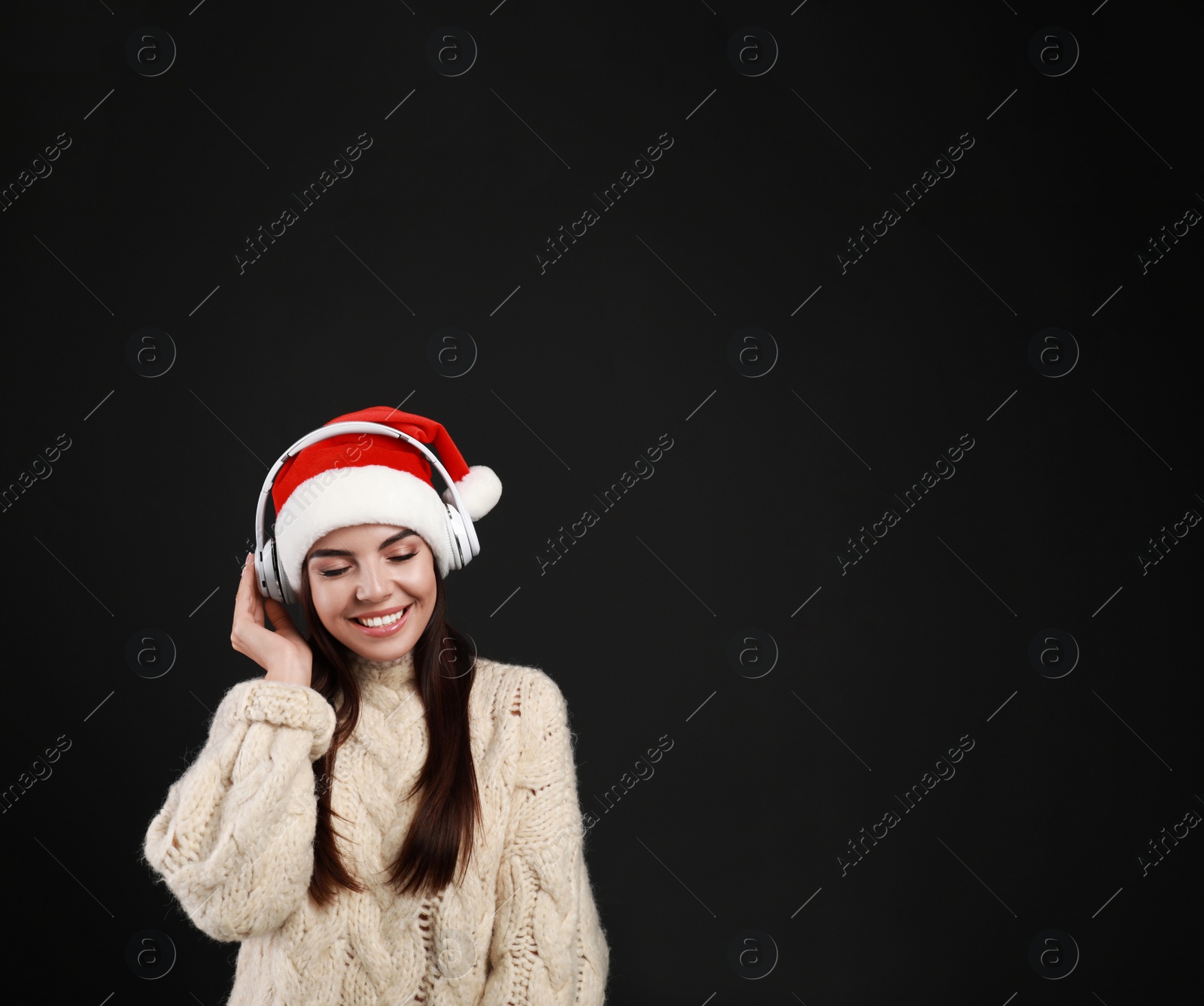 Photo of Young woman in Santa hat listening to Christmas music on black background, space for text