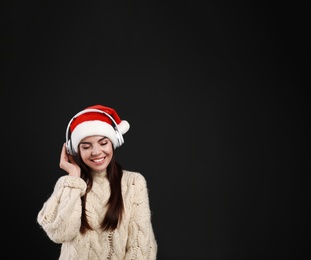 Photo of Young woman in Santa hat listening to Christmas music on black background, space for text