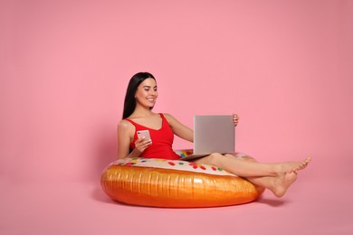 Photo of Young woman using laptop while sitting on inflatable ring against pink background