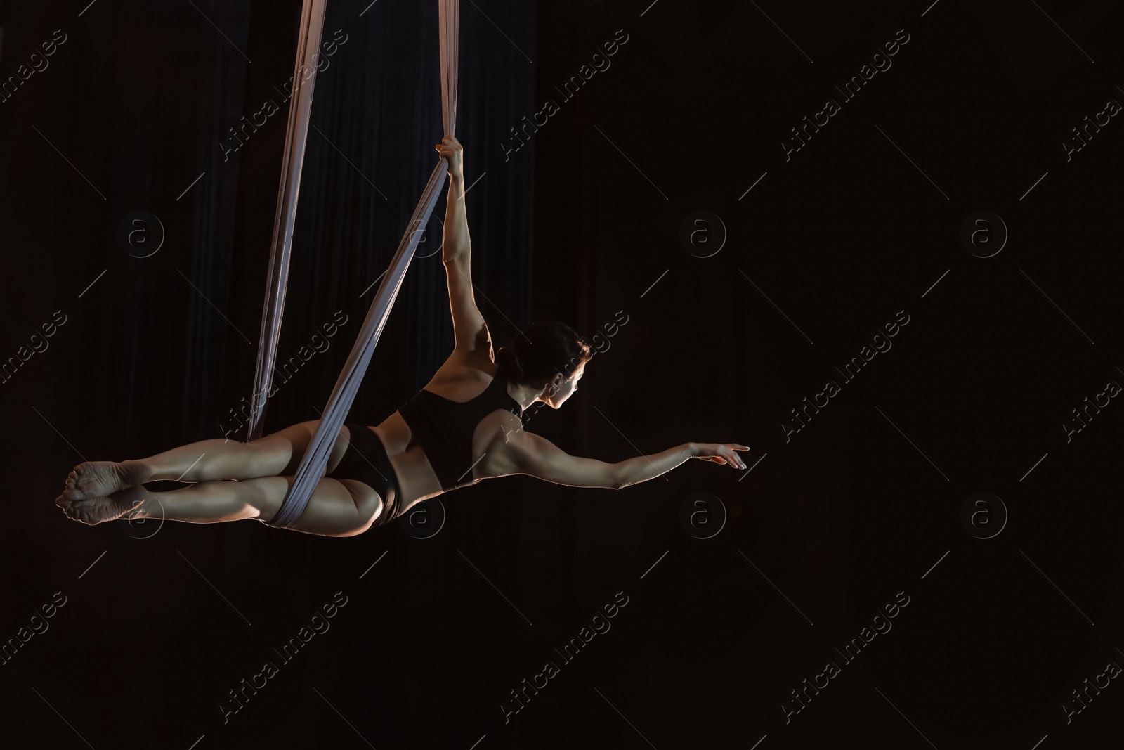 Photo of Young woman performing acrobatic element on aerial silk against dark background