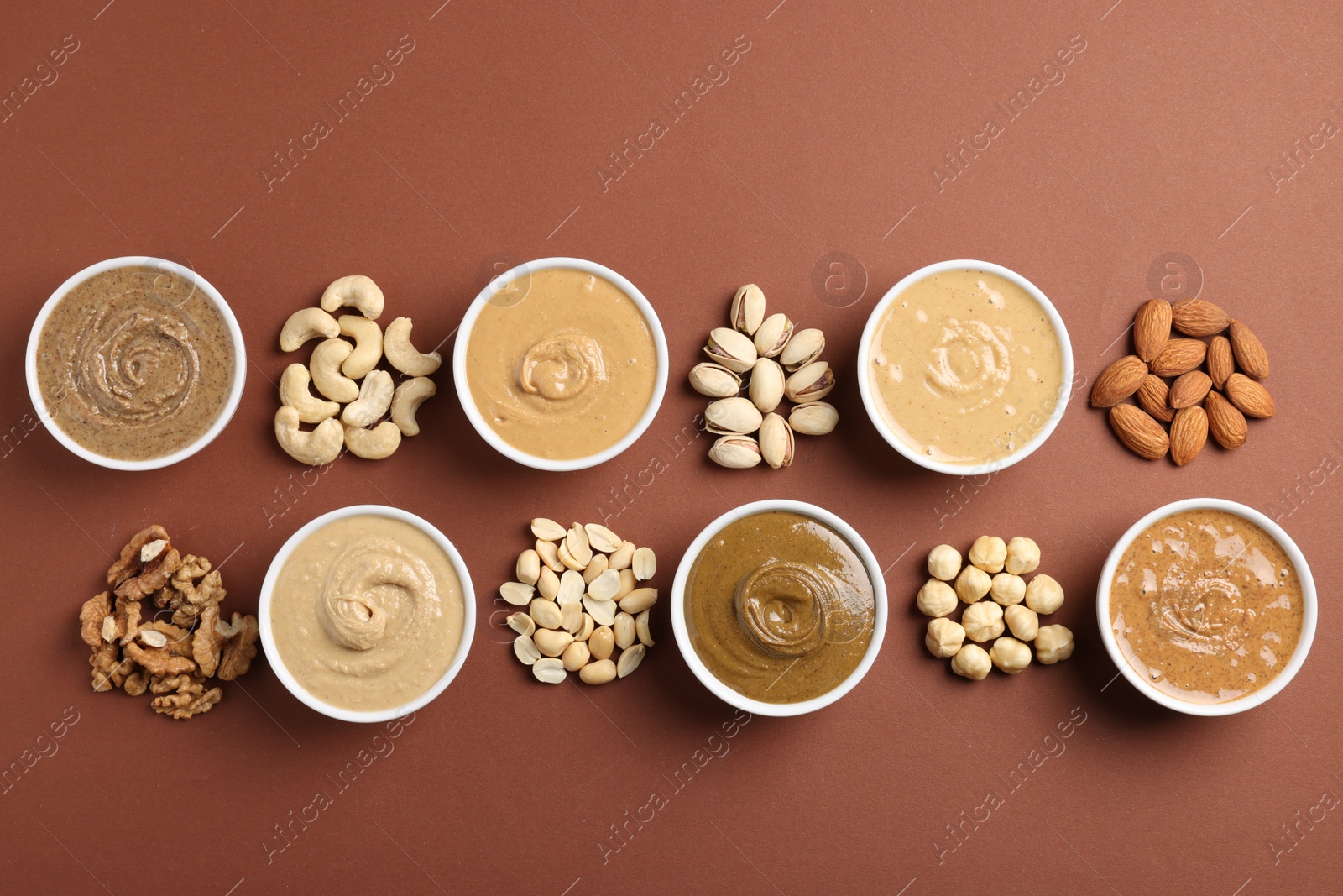 Photo of Many tasty nut butters in bowls and nuts on brown table, flat lay