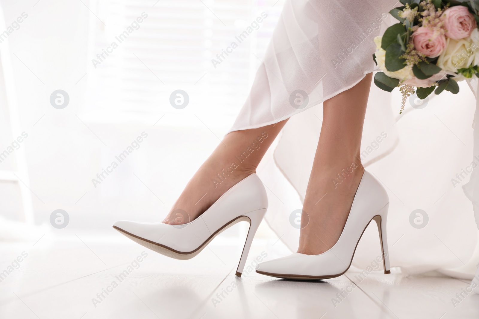Photo of Young bride with bouquet wearing wedding shoes indoors, closeup
