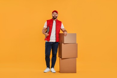Photo of Happy young courier with clipboard and stack of parcels on orange background