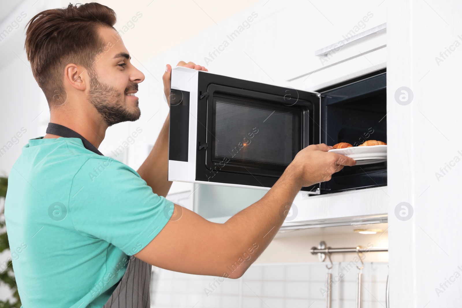 Photo of Young man putting plate with croissants into microwave oven at home