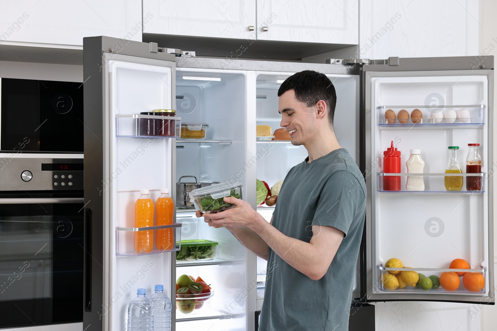 Photo of Happy man holding container with vegetables near refrigerator in kitchen