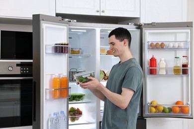 Photo of Happy man holding container with vegetables near refrigerator in kitchen