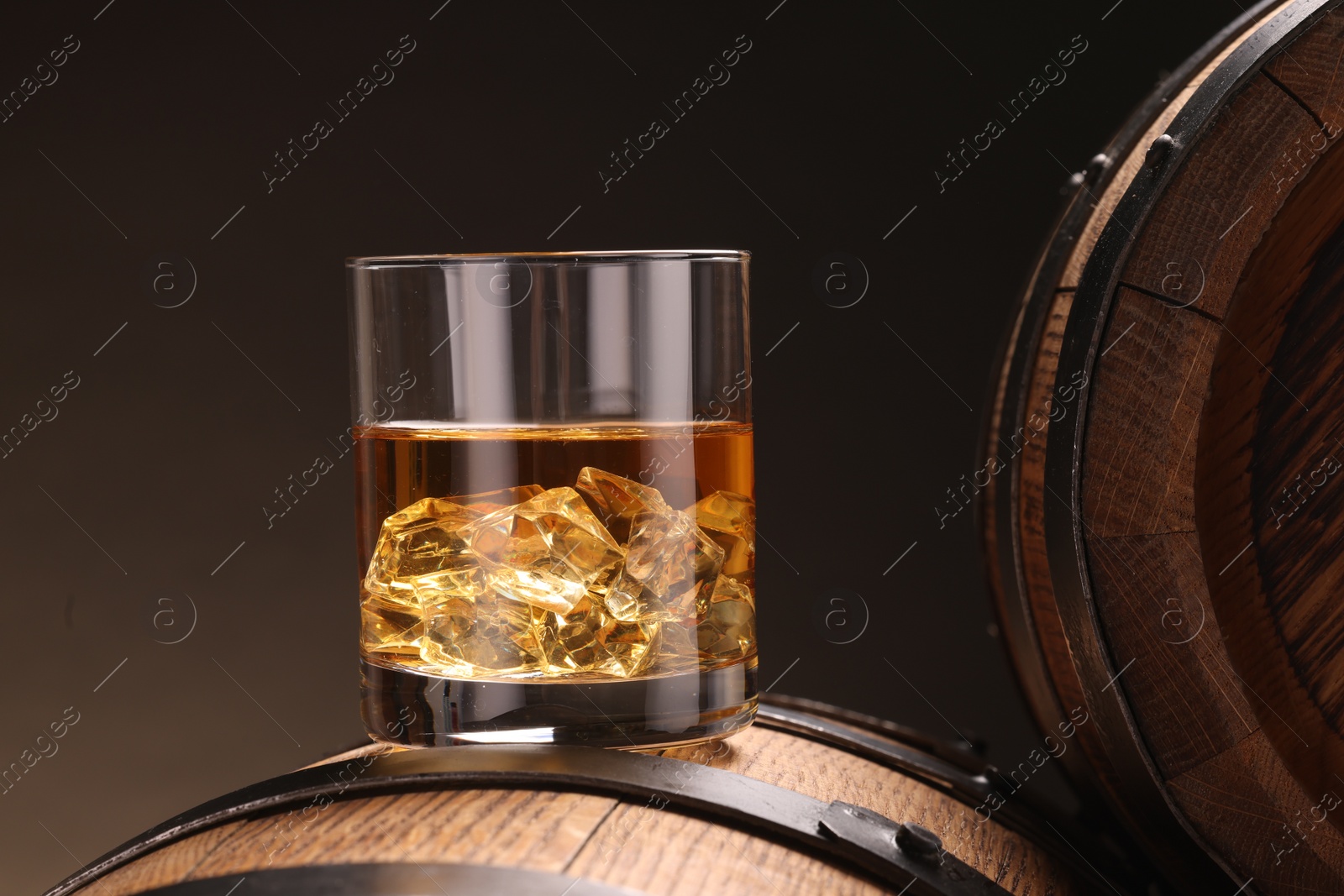 Photo of Whiskey with ice cubes in glass on wooden barrel against dark background, closeup