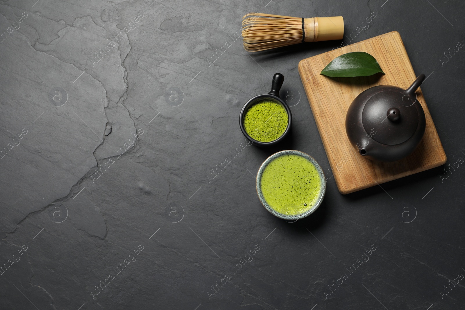 Photo of Cup of fresh matcha tea, bamboo whisk, teapot and green powder on black textured table, flat lay. Space for text