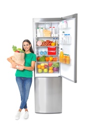 Young woman with bag of groceries near open refrigerator on white background