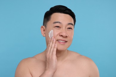 Happy man applying cream onto his face on light blue background