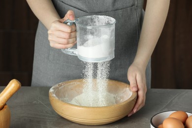 Photo of Woman sieving flour into bowl at grey table, closeup