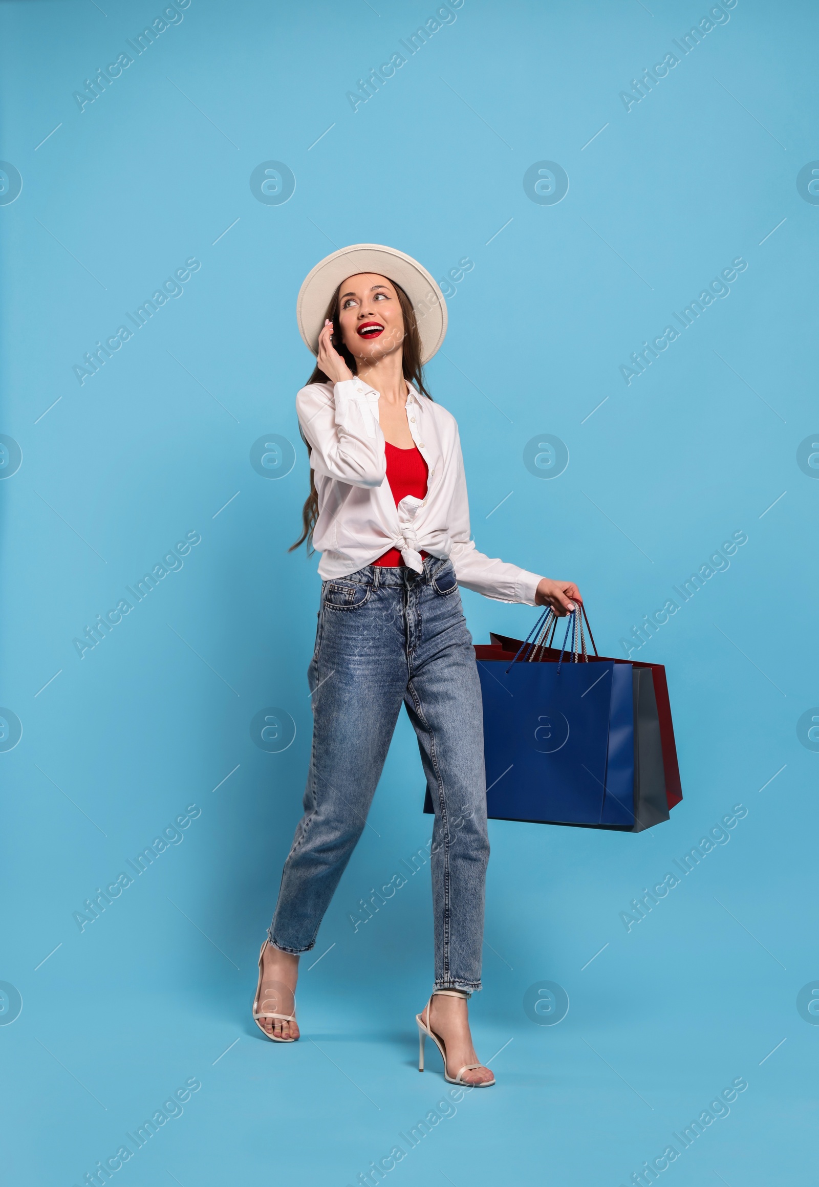 Photo of Stylish young woman with shopping bags talking on smartphone against light blue background