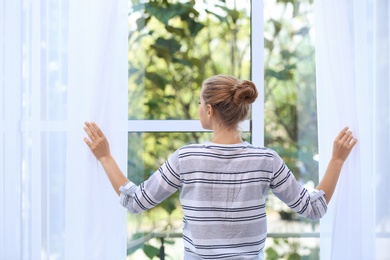 Photo of Young woman opening curtains and looking out of window at home