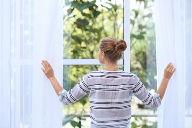 Young woman opening curtains and looking out of window at home