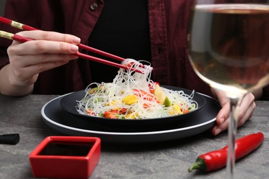 Photo of Stir-fry. Woman with chopsticks eating tasty rice noodles with meat and vegetables at grey textured table, closeup