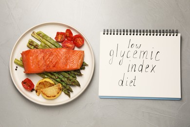 Photo of Notebook with words Low Glycemic Index Diet and plate of tasty grilled salmon on grey table, flat lay