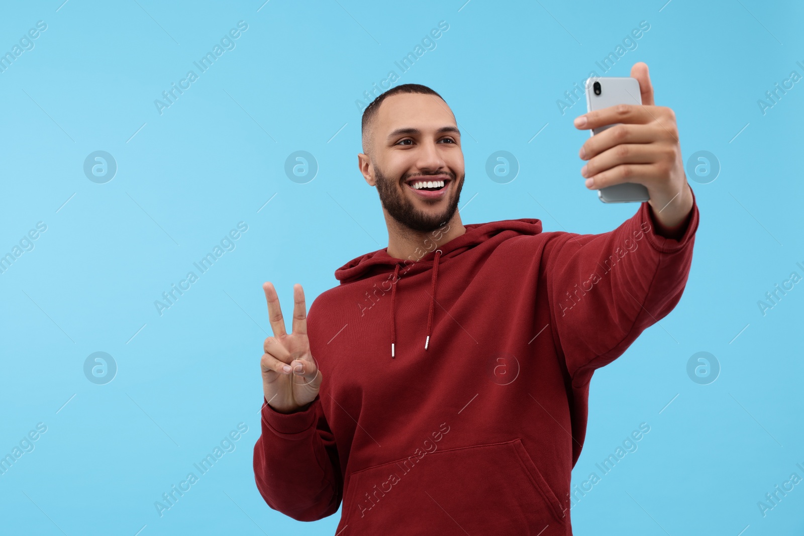 Photo of Smiling young man taking selfie with smartphone and showing peace sign on light blue background